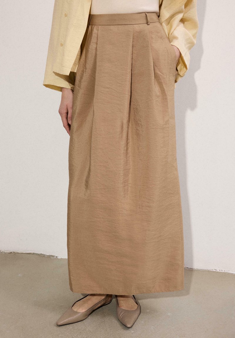 Μπεζ φούστα με ψηλή μέση έως τον αστράγαλο με πτυχώσεις μπροστά, συνδυασμένη με μπεζ μυτερές μπαλαρίνες και ανοιχτό κίτρινο μακρυμάνικο τοπ, με τα χέρια στις τσέπες.