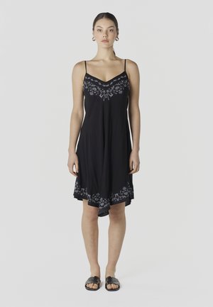 Mujer con un vestido negro hasta la rodilla con tirantes finos y bordado floral en el pecho y el dobladillo, combinado con sandalias negras tipo slide.