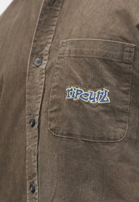 Camisa de bombazine castanha com botões pretos, apresentando um bolso no lado esquerdo do peito e o logótipo "Rip Curl" bordado em azul e amarelo.