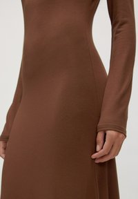 Nahaufnahme einer Person in einem eng anliegenden, langärmligen braunen Kleid, die die Körpermitte zeigt und eine Hand entspannt an der Seite hat.