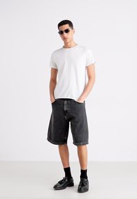 Weißes kurzärmliges T-Shirt kombiniert mit schwarzen Jeansshorts und schwarzen Schuhen mit Quasten. Das Model trägt dunkle Sonnenbrillen.
