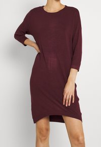 Donna che indossa un vestito bordeaux a maniche lunghe e lunghezza al ginocchio, in piedi con una mano sulla vita.