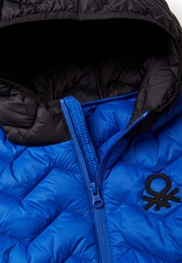 Chaqueta acolchada azul con capucha negra, que presenta una cremallera azul y un logo bordado negro en el pecho. Tejido suave y ligero.