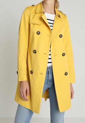 Kvinde iført en skarp gul dobbeltradet trenchcoat med sorte knapper over en stribet skjorte og lyseblå jeans.