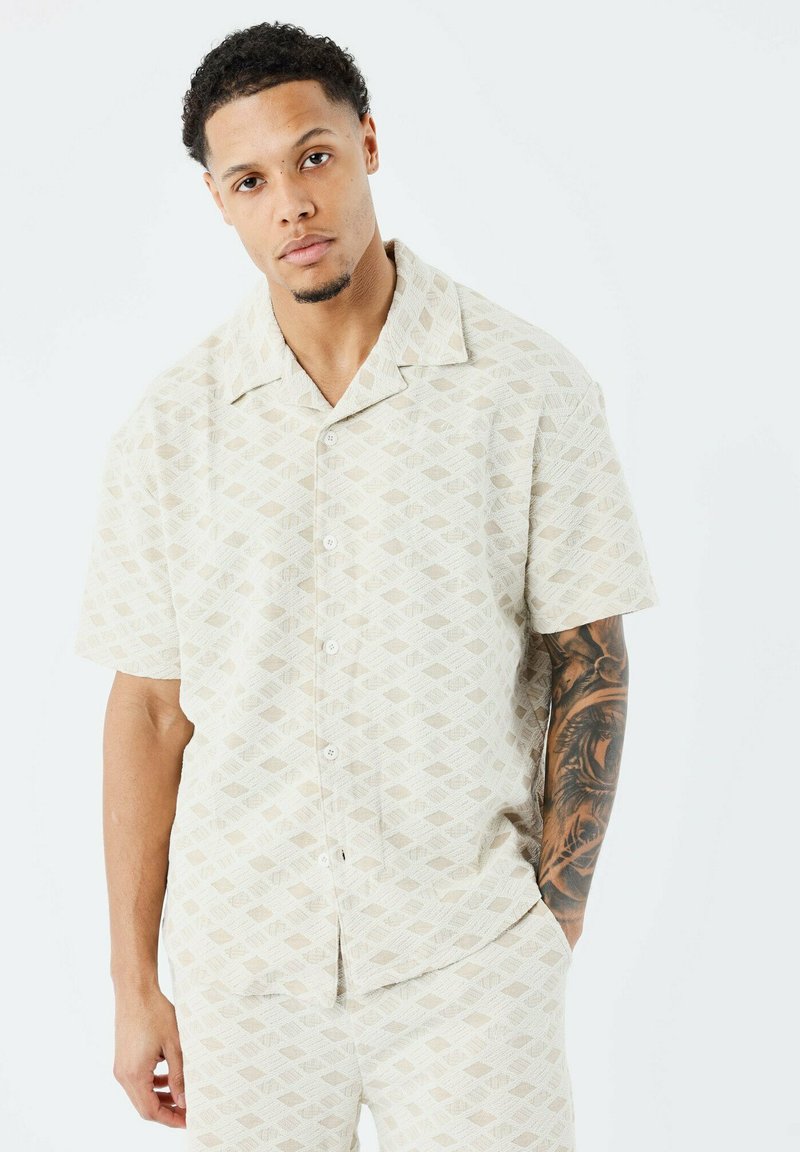 Camisa de manga corta y botones hecha de una tela beige con un patrón de formas de diamante. El cuello es ancho y tiene un corte relajado.