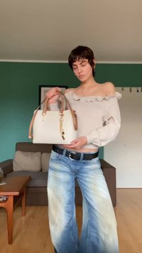 Bolso blanco con asas de cuero marrón, herrajes dorados y un acabado texturizado, sostenido por una persona que lleva un suéter de volantes gris claro y jeans.