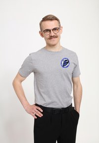Grå kortærmet t-shirt med rund udringning og et blåt emblem på venstre bryst, parret med sorte bukser og et sort bælte.