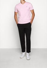 T-shirt a V rosa in tessuto morbido, abbinata a pantaloni slim-fit neri. Sneakers bianche completano l'outfit. Design semplice, senza motivi visibili.