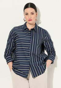 Camicia blu navy con righe verticali color crema, design con bottoni, colletto, taschino sul petto e maniche arrotolate. Tessuto con texture liscia.