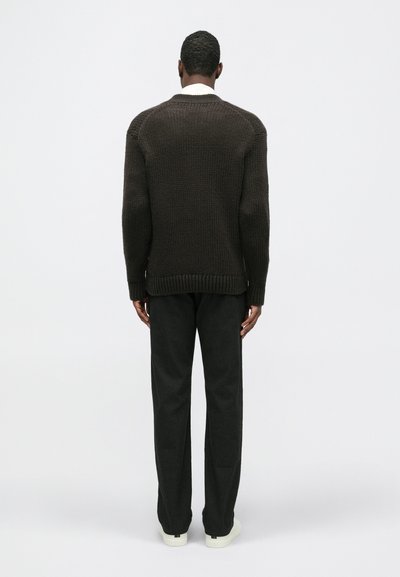 Sötétbarna kötött pulóver bordás szegéllyel és mandzsettával, sötét nadrággal és fehér tornacipővel párosítva, hátulról nézve, világos háttér előtt.