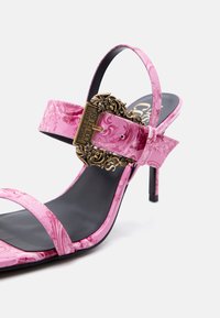 Ροζ γυναικείο πέδιλο με χοντρό τακούνι, μαρμάρινο μοτίβο, λεπτά λουριά, μαύρη σόλα και περίτεχνο χρυσό αγκράφα Versace Jeans Couture.