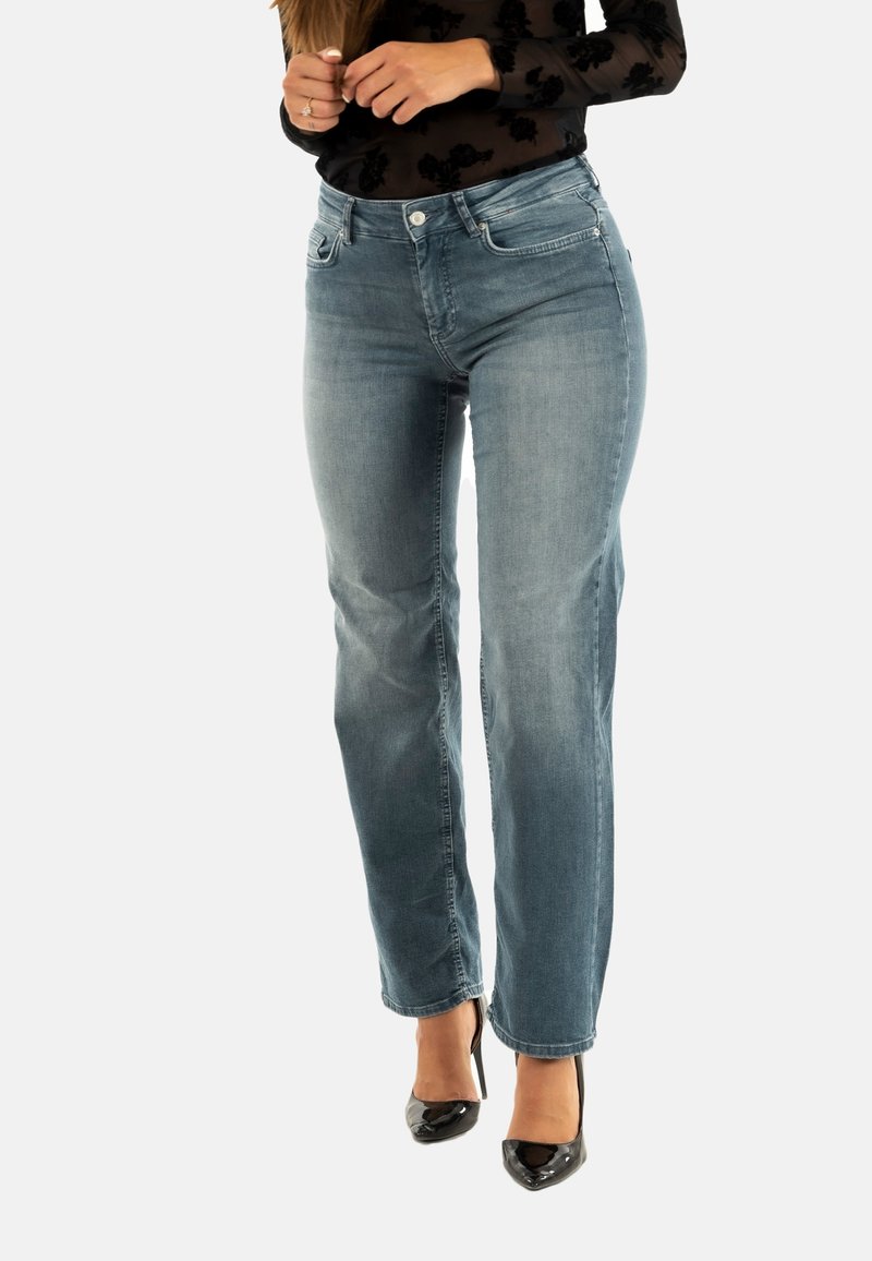 Donna che indossa jeans dritti blu sbiaditi e tacchi alti neri a punta, mani incrociate vicino alla vita, su uno sfondo semplice.