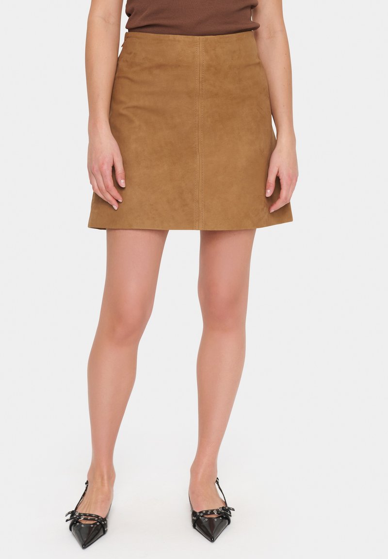 Jupe mini en daim marron avec une coupe droite, des détails de couture au centre et une texture lisse. Associée à des escarpins noirs à bout pointu.