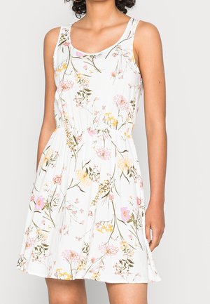 Vrouw draagt een mouwloze witte jurk met een bloemmotief in roze, geel en groen, met een geplooide taille en zakken.