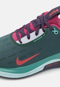 Žalios ir mėlynos sportinės bateliai su rožiniais akcentais, tekstūruota viršutinė dalis, violetinės raišteliai ir raudonas Nike logotipas. Ypatybės: kvėpuojanti medžiaga ir neslystantis padas.