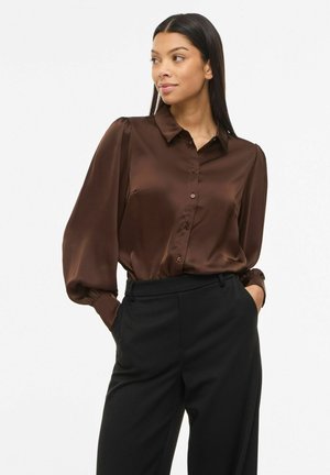 Femme aux longs cheveux noirs portant un chemisier marron brillant à boutons avec des manches bouffantes et un pantalon noir taille haute, mains dans les poches, regardant vers la droite.