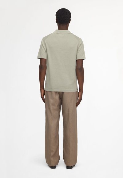 Uomo visto di spalle che indossa una polo a maniche corte grigio chiaro e pantaloni marroni larghi, davanti a uno sfondo bianco semplice.