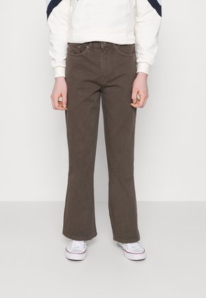 Person trägt braune, gerade geschnittene Hose, ein weißes Sweatshirt mit dunkelblauen Streifen an den Ärmeln und weiße Turnschuhe, steht vor einem einfarbigen Hintergrund.