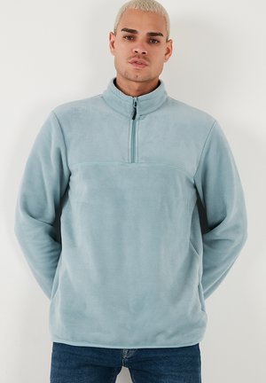 Lys blå fleece trøje med høj krave og halv lynlås foran. Blød tekstur med et ensartet design og en vandret søm på tværs af brystet.