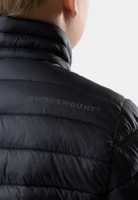 Sort pufferjakke med quiltet design og høj krave. "SWEDEMOUNT" logo trykt på ryggen i en diskret grå tone.