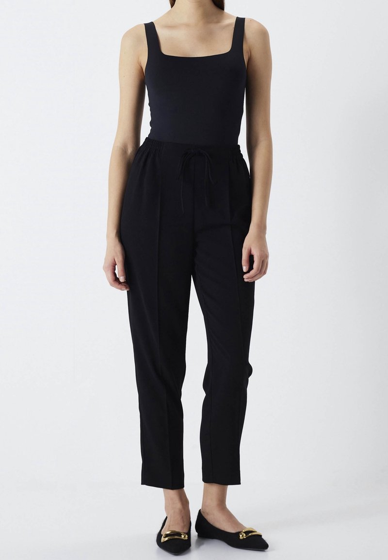 Femme portant un haut noir sans manches à encolure carrée, un pantalon noir taille haute avec cordon de serrage, et des ballerines noires avec un détail de boucle dorée.