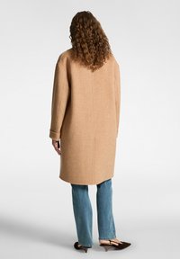 Cappotto in lana color cammello con una vestibilità ampia, maniche lunghe e taglio fino a metà coscia. Indossato sopra jeans azzurri chiari e scarpe slip-on scure.