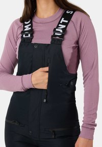 Pettorine nere con bretelle regolabili, dotate di logo, tasche frontali e aperture con cerniera. Indossate sopra una maglietta a coste di un leggero color viola.