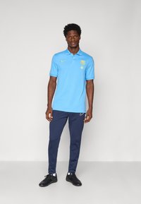 Svetlo modra polo majica s ovratnikom, rumenim logotipom, v kombinaciji z mornarskimi atletskimi hlačami in črnimi čevlji. Gladek material in prilagojen kroj.