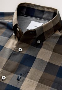 Brown and navy plaid shirt with a button-down collar, fabricated from a smooth cotton blend. Features white buttons and a small logo patch.