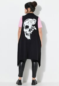 Schwarzes ärmelloses Oberteil mit einem großen weißen Totenkopf- und Rosenmotiv auf dem Rücken, getragen über einem pinken Shirt und schwarzen Lederleggings.