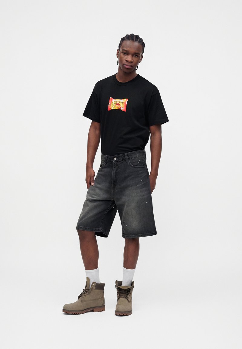 Man with braided hair wearing a black graphic t-shirt, faded black denim shorts, white socks, and beige lace-up boots, standing against white background.