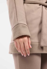 Giacca beige con tessuto liscio e polsini in pelliccia texture. Le maniche terminano con un orlo piegato, mostrando un design senza cuciture.