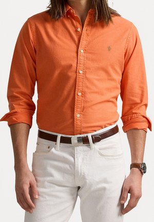 Homme portant une chemise orange à manches longues boutonnée, manches retroussées, pantalon blanc, ceinture marron et une montre-bracelet au poignet gauche.