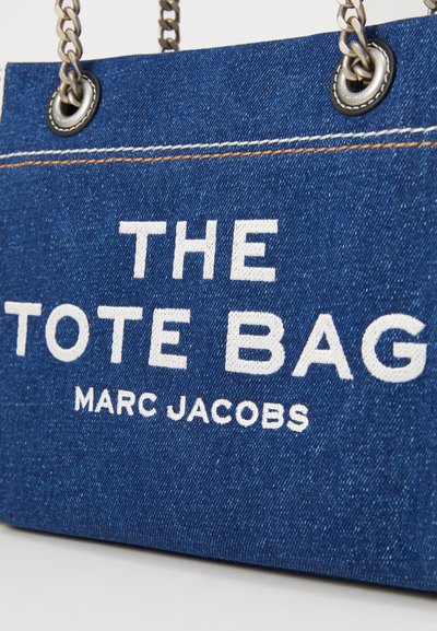 Denim iepirkumu soma ar sudraba ķēdes rokturiem. Priekšpusē liela balta teksta uzraksts "THE TOTE BAG" un "MARC JACOBS" uz zila teksturēta fona.