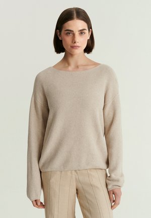 Vrouw met kort bruin haar, die een beige gebreide trui met textuur draagt en lichtbeige broek met verticale strepen, staand tegen een effen achtergrond.