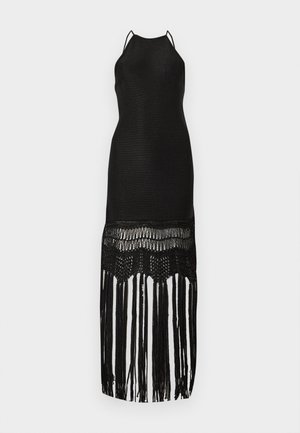 Kvinde iført en ærmeløs sort kjole med frynsekant, holder en cremefarvet clutch taske og har hvide høje hæle sandaler på.