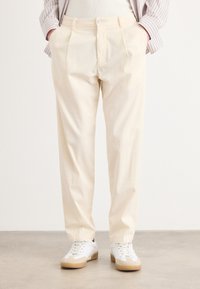 Pantalones plisados de color beige claro, hechos de algodón, con corte recto y bolsillos laterales, combinados con zapatillas blancas y una camiseta a rayas.