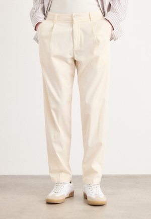 Pleated light beige trousers made from cotton, featuring a straight cut and side pockets, paired with white sneakers and a striped shirt.