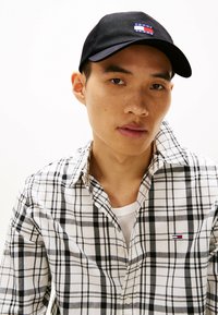 Sort baseball cap med logo, hvid skjorte med sort ternet mønster, lagdelt over en hvid undertrøje, synlige knapper og krave detaljer.