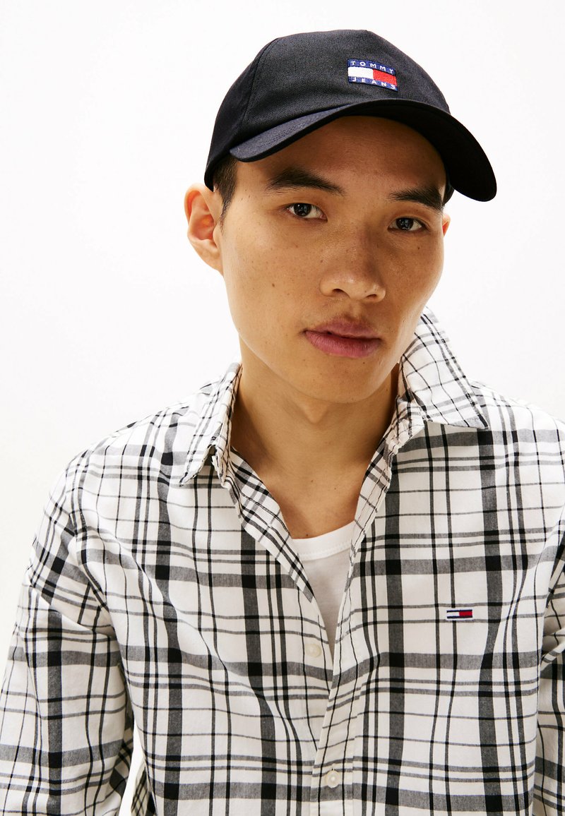 Sort baseball cap med logo, hvid skjorte med sort ternet mønster, lagdelt over en hvid undertrøje, synlige knapper og krave detaljer.