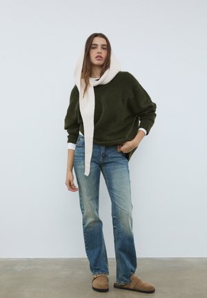 Dark green oversized sweater with a textured finish, paired with light blue straight-leg jeans and tan slip-on shoes. White ribbed scarf drapes around the neck.