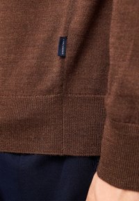 Maglione lavorato a maglia marrone con texture a coste e un'etichetta di marca visibile. Presenta maniche lunghe e una vestibilità rilassata. Viene abbinato a pantaloni blu scuro.