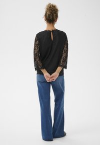 Sort blonde skjorte med trekvart-lange ærmer og nøglehulslukning i ryggen, parret med lyseblå flagrende jeans. Glat tekstur, bølget kant.