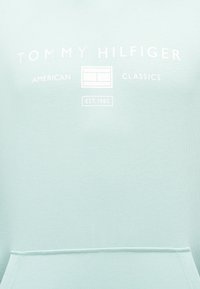 Λογότυπο Tommy Hilfiger με το κείμενο "American Classics", το έμβλημα της μάρκας και τη φράση "EST. 1985" σε ανοιχτό γαλάζιο ύφασμα πάνω από μια τσέπη καγκουρό.