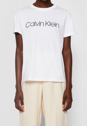 Osoba nosząca białą koszulkę z krótkim rękawem z czarnym tekstem "Calvin Klein" i jasnobeżowymi spodniami na jednolitym tle.
