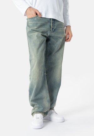Person iført falmede, løstsiddende blå jeans, hvid langærmet skjorte og hvide sneakers, med den ene hånd i lommen mod en ensfarvet baggrund.