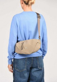 Beige crossbodytas van zacht materiaal, met een afgeronde vorm, een enkele bruine band en een ritssluiting. Logo zichtbaar aan de voorkant.