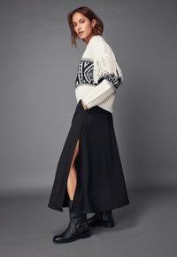 Pull créme épais avec un motif géométrique noir et des franges, associé à une longue jupe noire avec une fente sur le côté, porté avec des bottes noires.