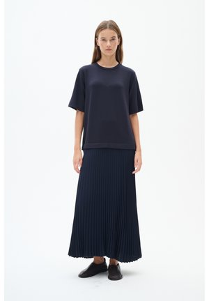Žena s dlhými rovnými vlasmi, oblečená v voľnom tmavomodrom tričku s krátkym rukávom, plisovanej tmavomodrej maxisukni a čiernych slip-on topánkach, stojaca pred bielym pozadím.