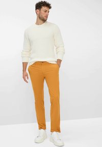 Witte gebreide trui met textuur, gecombineerd met feloranje slim-fit broek en witte sneakers, tentoon gesteld op een mannelijke model.